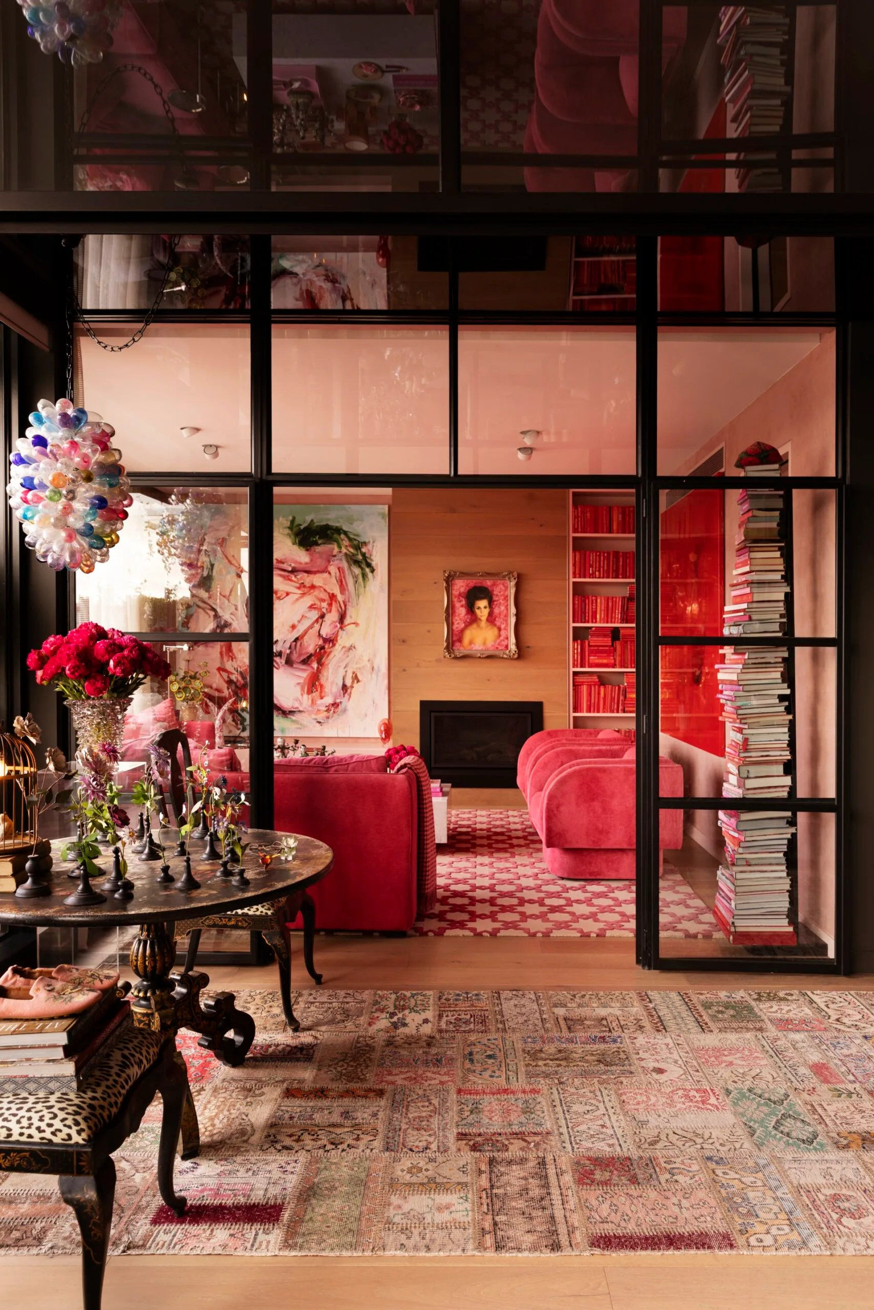 Opulent interior with pink sofas, art, books, floral decor, colorful chandelier, and patterned rug in a cozy room.