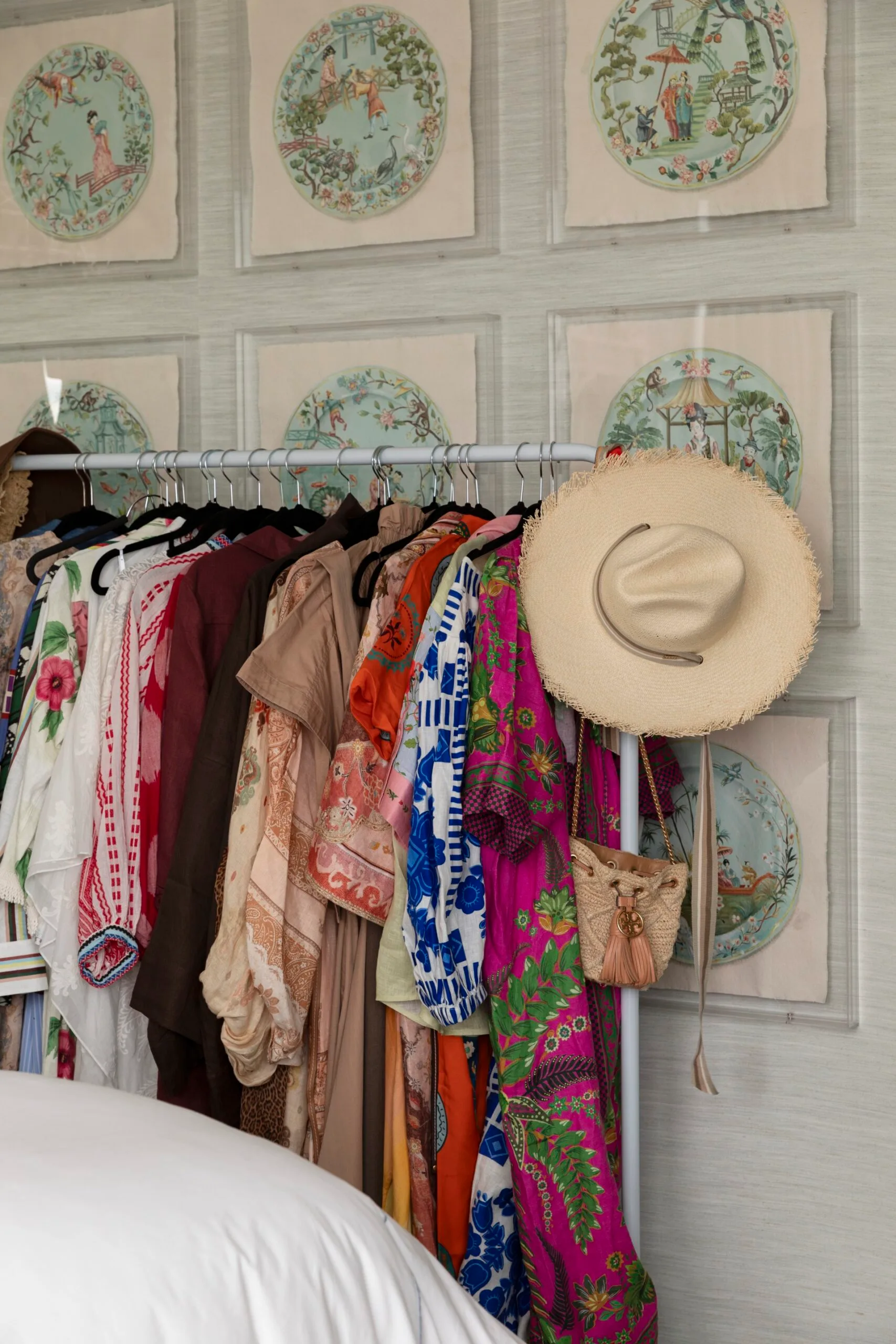 Colorful clothes on a rack with a hat and purse; decorative Asian art on walls; bedroom setting.