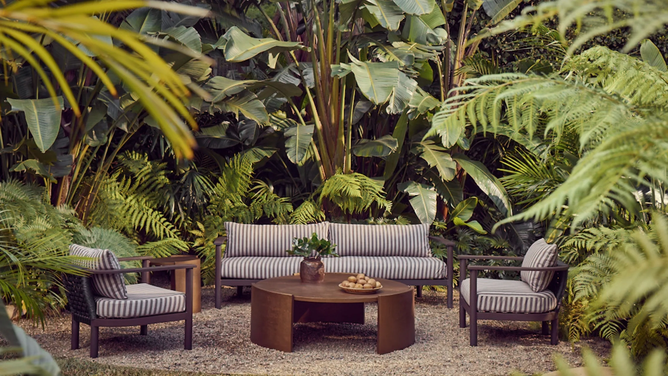 Outdoor seating set with striped cushions in a lush garden surrounded by ferns and tropical plants.