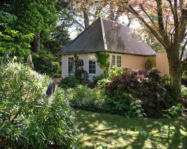 Cottage with a sloped metal roof surrounded by lush greenery and trees in a garden setting.