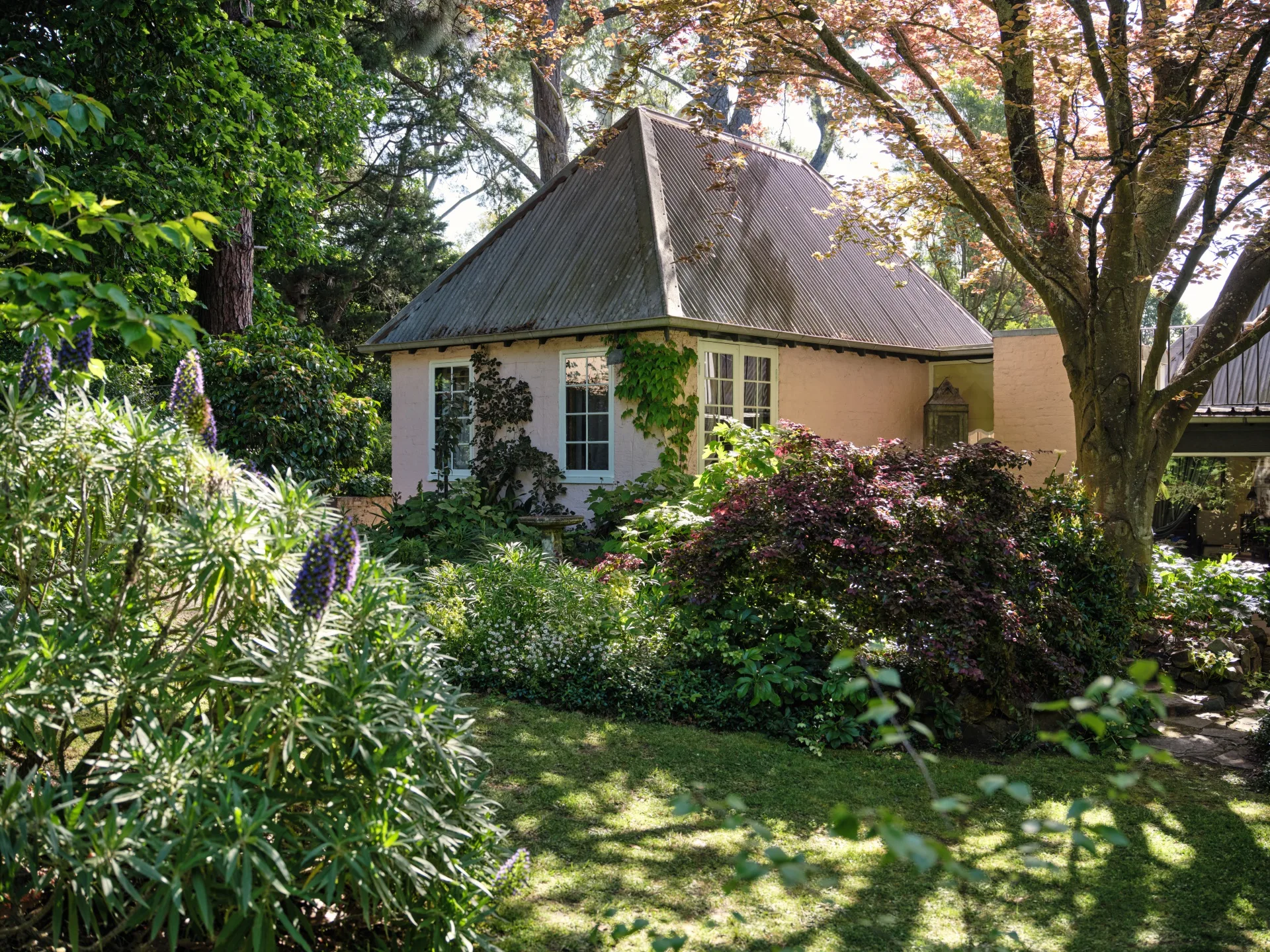 Cottage with a sloped metal roof surrounded by lush greenery and trees in a garden setting.