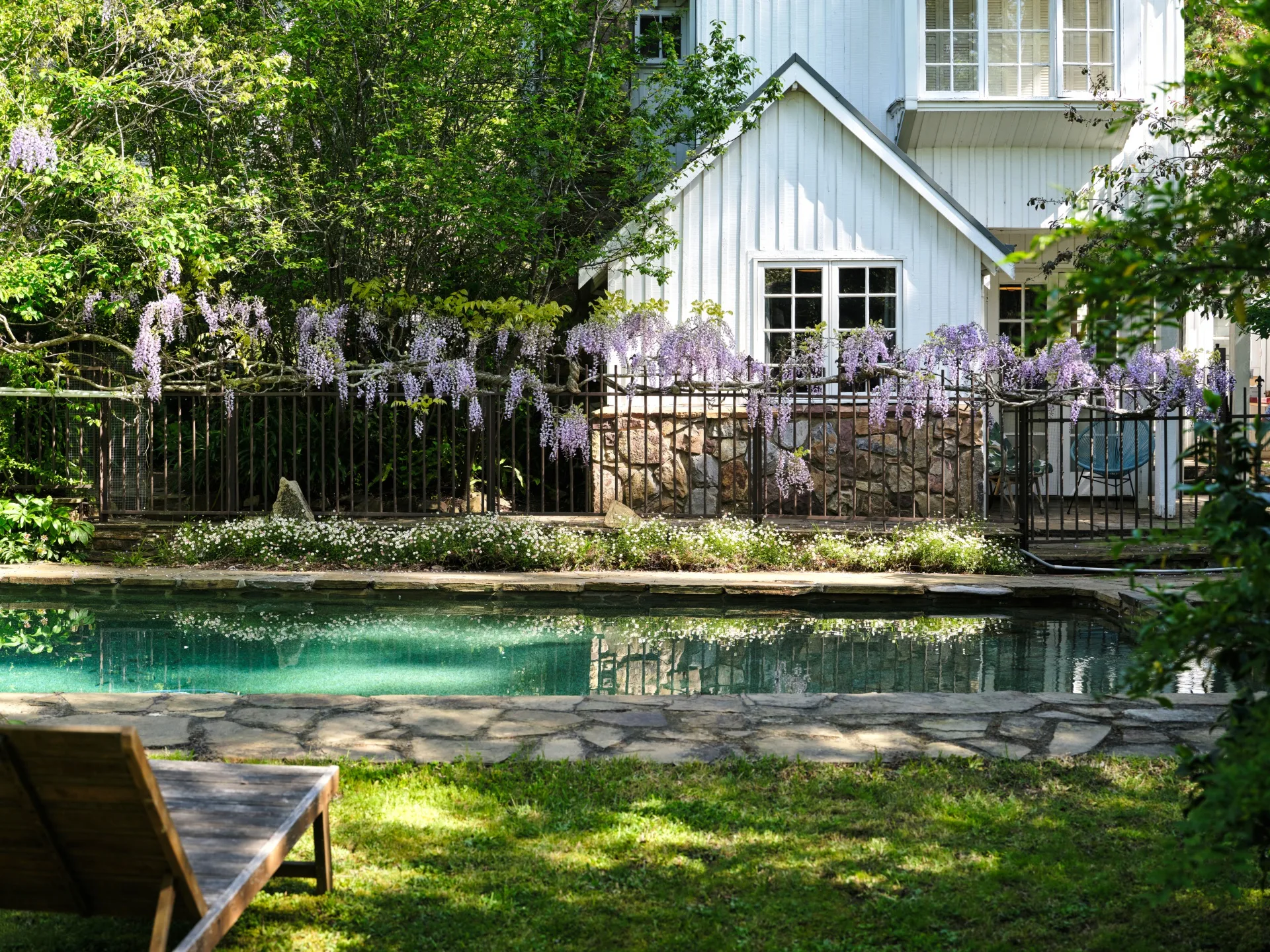White house with wisteria-covered fence beside a pool and wooden lounge chair on a sunny day. This is part of Bickleigh Village, created by Edna Walling.