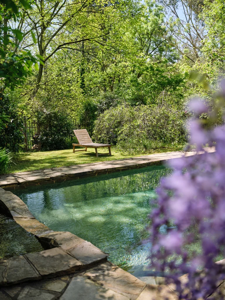 Stone-lined pool with a wooden lounge chair, surrounded by lush greenery and purple wisteria in the foreground. This is part of Bickleigh Village, created by Edna Walling.