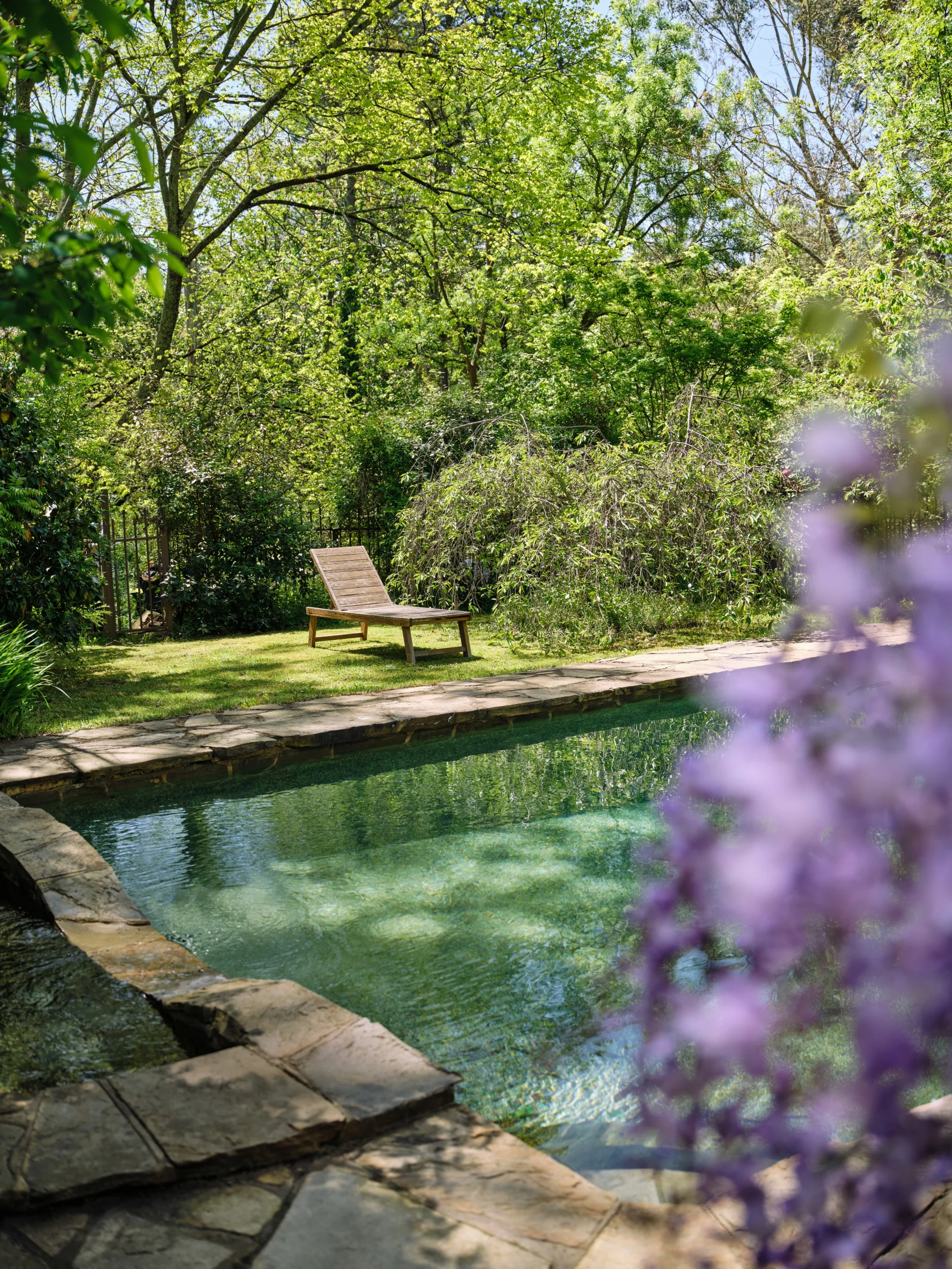 Stone-lined pool with a wooden lounge chair, surrounded by lush greenery and purple wisteria in the foreground. This is part of Bickleigh Village, created by Edna Walling.