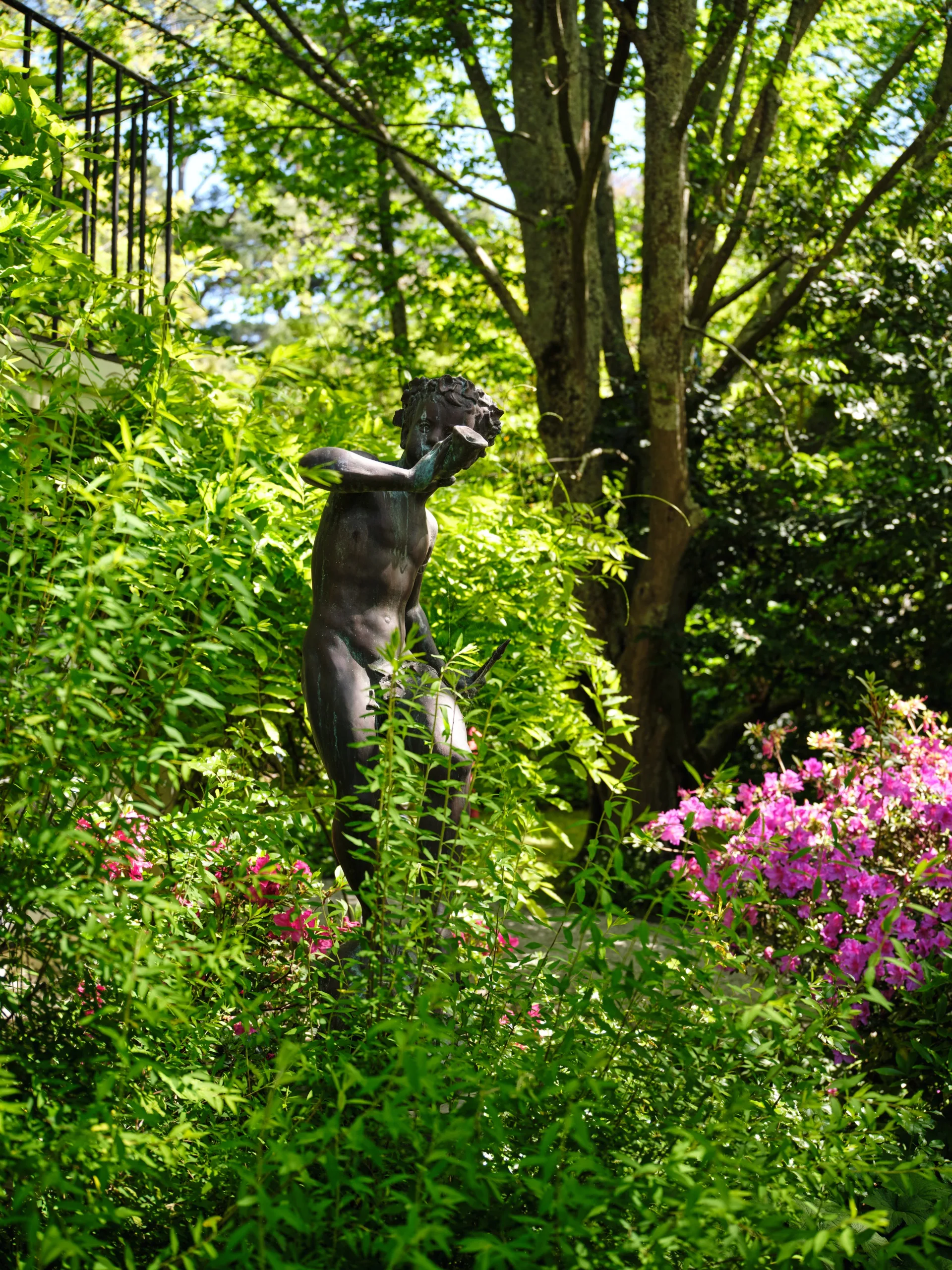 Statue of Pan playing a flute amidst lush greenery and pink flowers in a garden setting