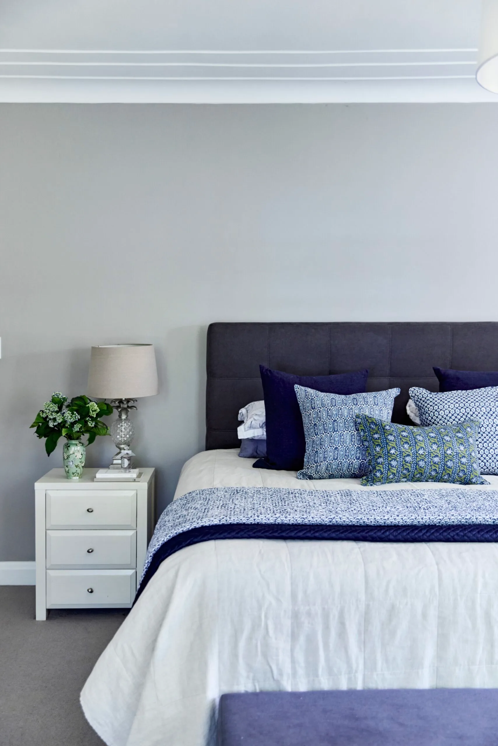 Bedroom with gray and blue bedding, a dark grey bedhead, white side table with lamp, books, and a floral vase.