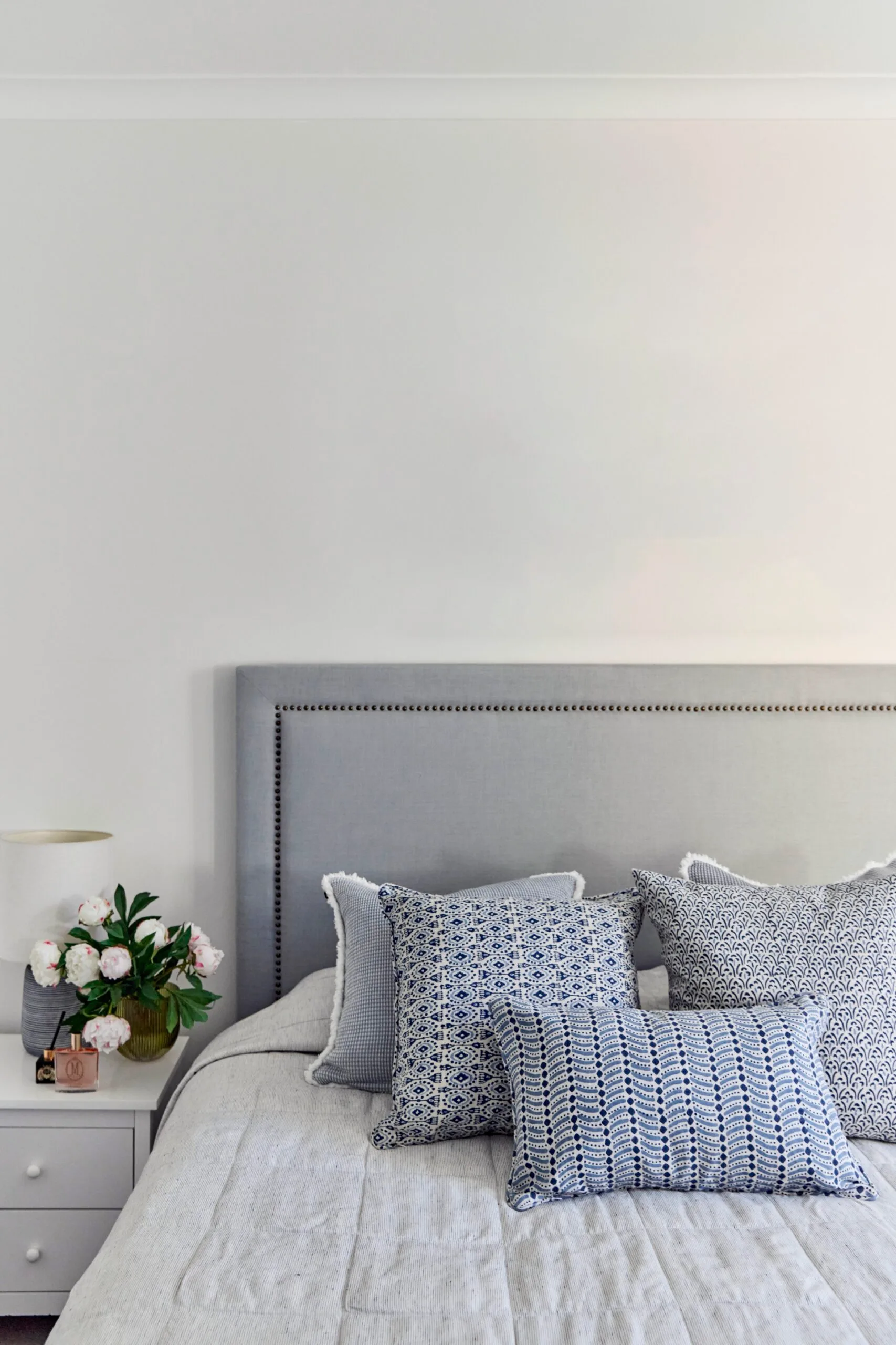Cozy bedroom with a grey headboard, patterned blue and white cushions, and a bedside table with a lamp and flowers.