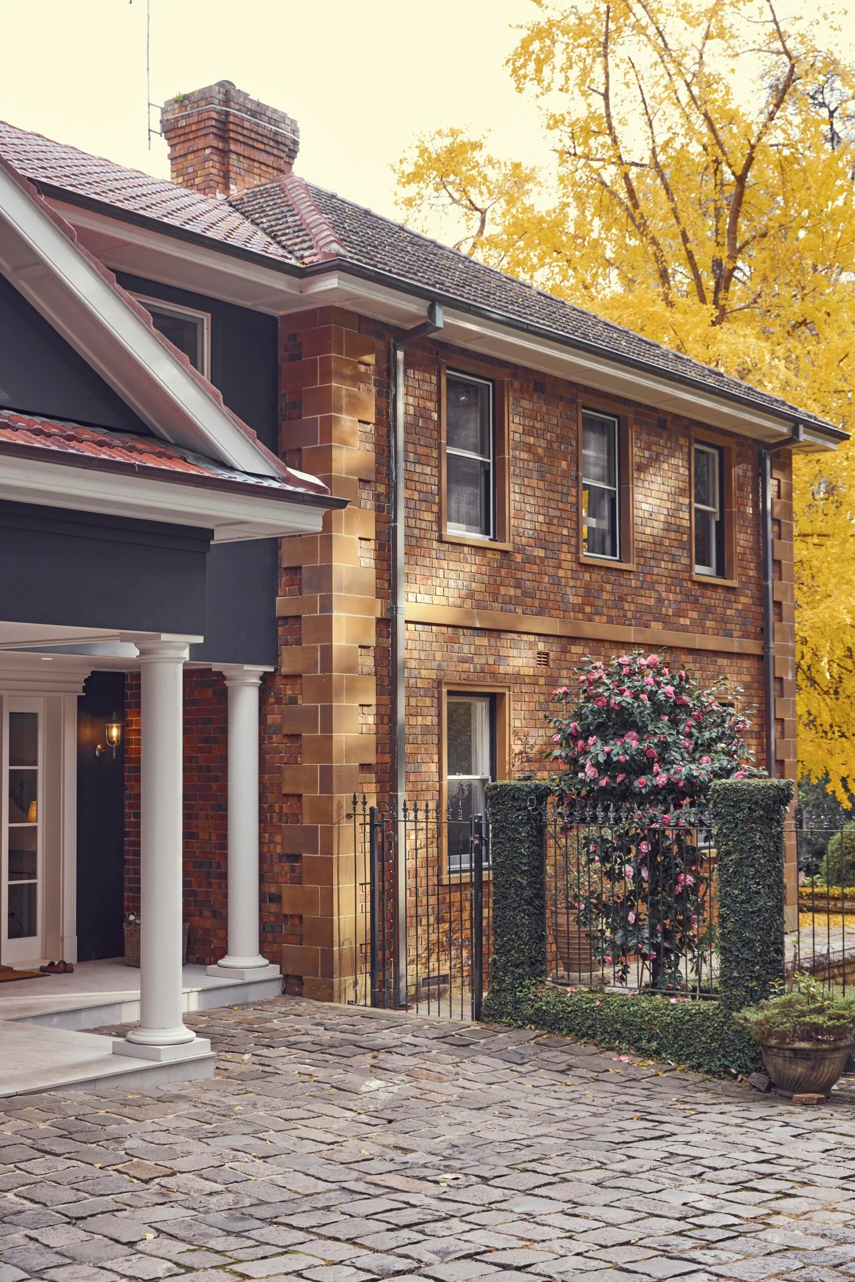 Historic brick house with white columns, gated entrance leading to a pool, and pink flowering shrub. Stone pathway and autumn trees in background. The facade of this heritage house is a Georgian style.