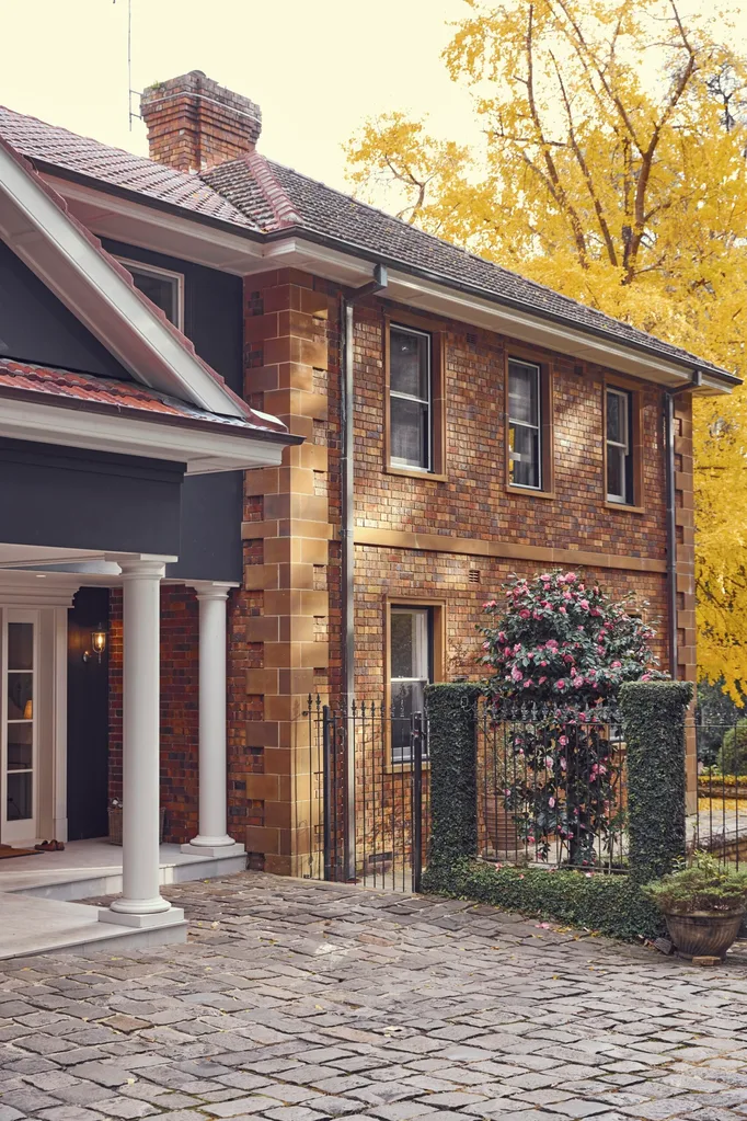 Historic brick house with white columns, gated entrance leading to a pool, and pink flowering shrub. Stone pathway and autumn trees in background. The facade of this heritage house is a Georgian style.