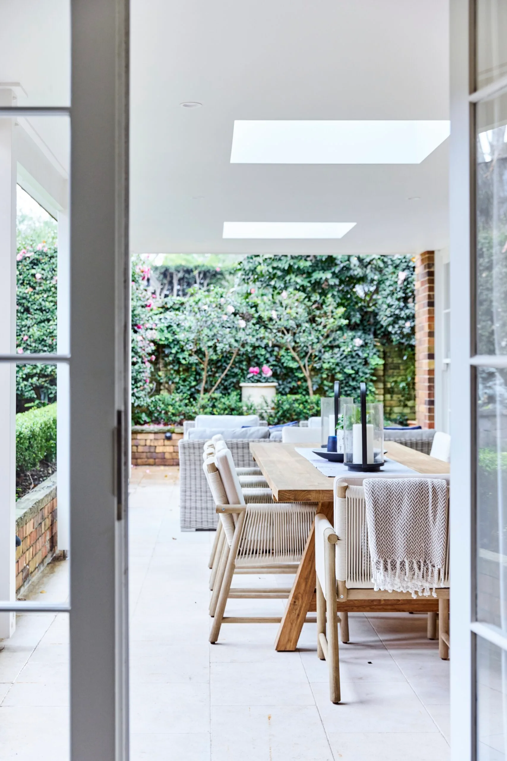 The outdoor patio of a heritage house with an alfresco dining area with an alfresco living area in the background. There is a wooden dining table, chairs, and candle holders, overlooking a garden with lush greenery and flowers.