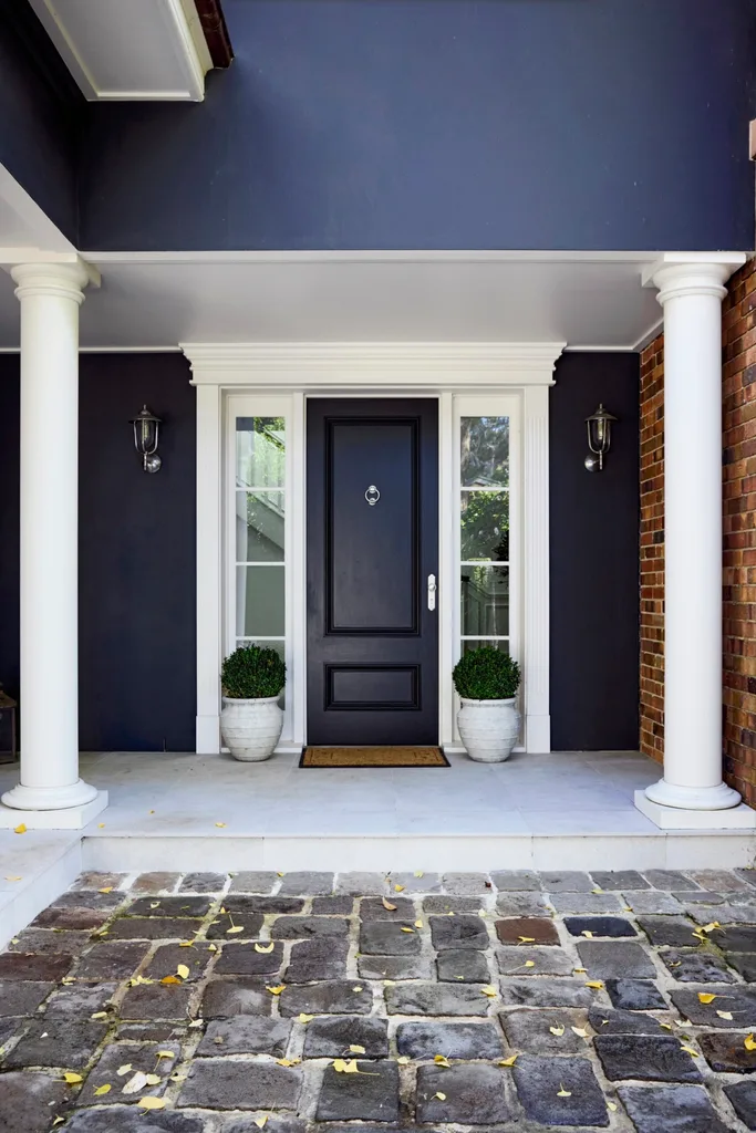 Front porch of a heritage house with black door, white frame and columns, two potted plants, and stone paving.