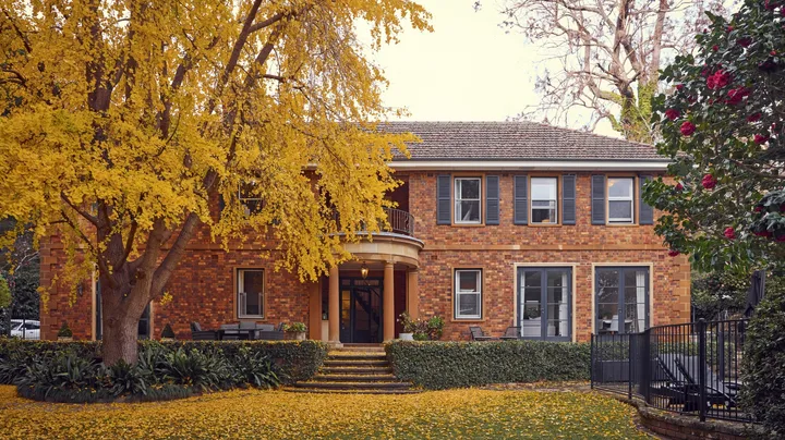 Red-brick house with a large yellow tree in front, surrounded by a garden with fallen leaves. The facade of this heritage house is a classic Georgian style.