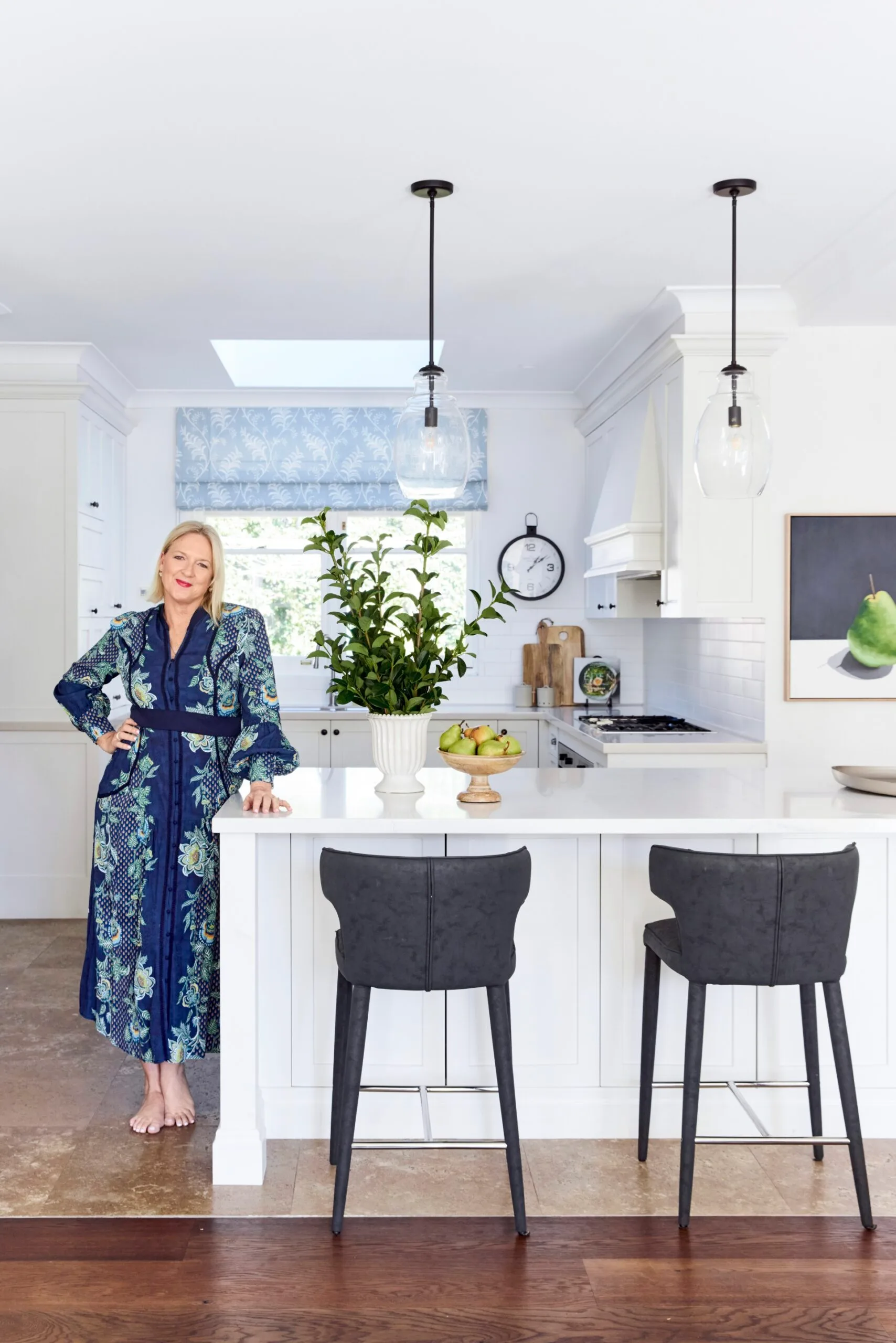 Woman in a blue patterned dress stands barefoot in a bright kitchen with white cabinets and a central island. A blue Roman blind is on the window, with two pendant lights hanging above the island. The rangehood has a traditional shape, suiting the heritage house.