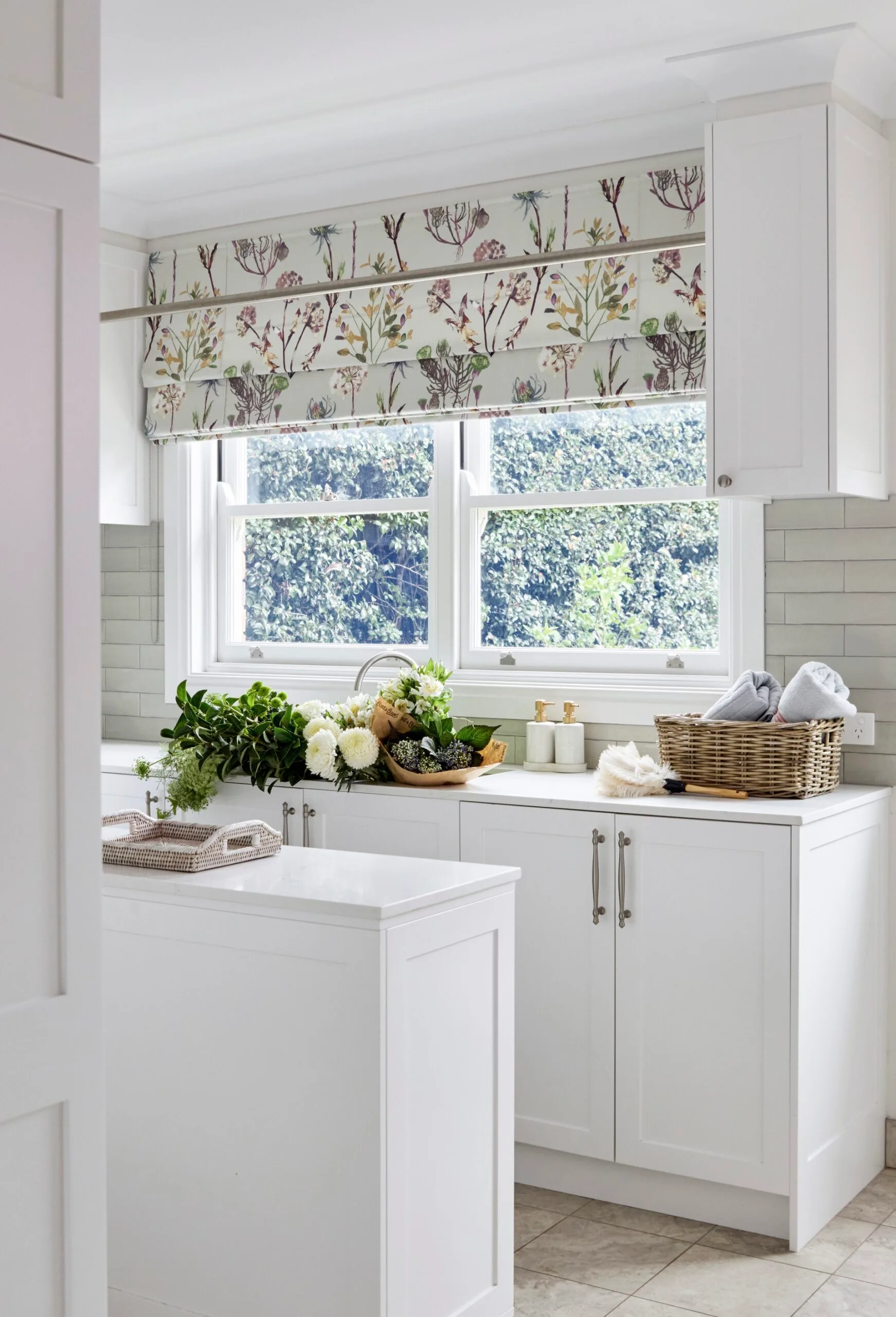 Bright laundry with white cabinets, floral blinds, and a basket with folded towels on the counter. Greenery and cut flowers have been placed in the large laundry sink.
