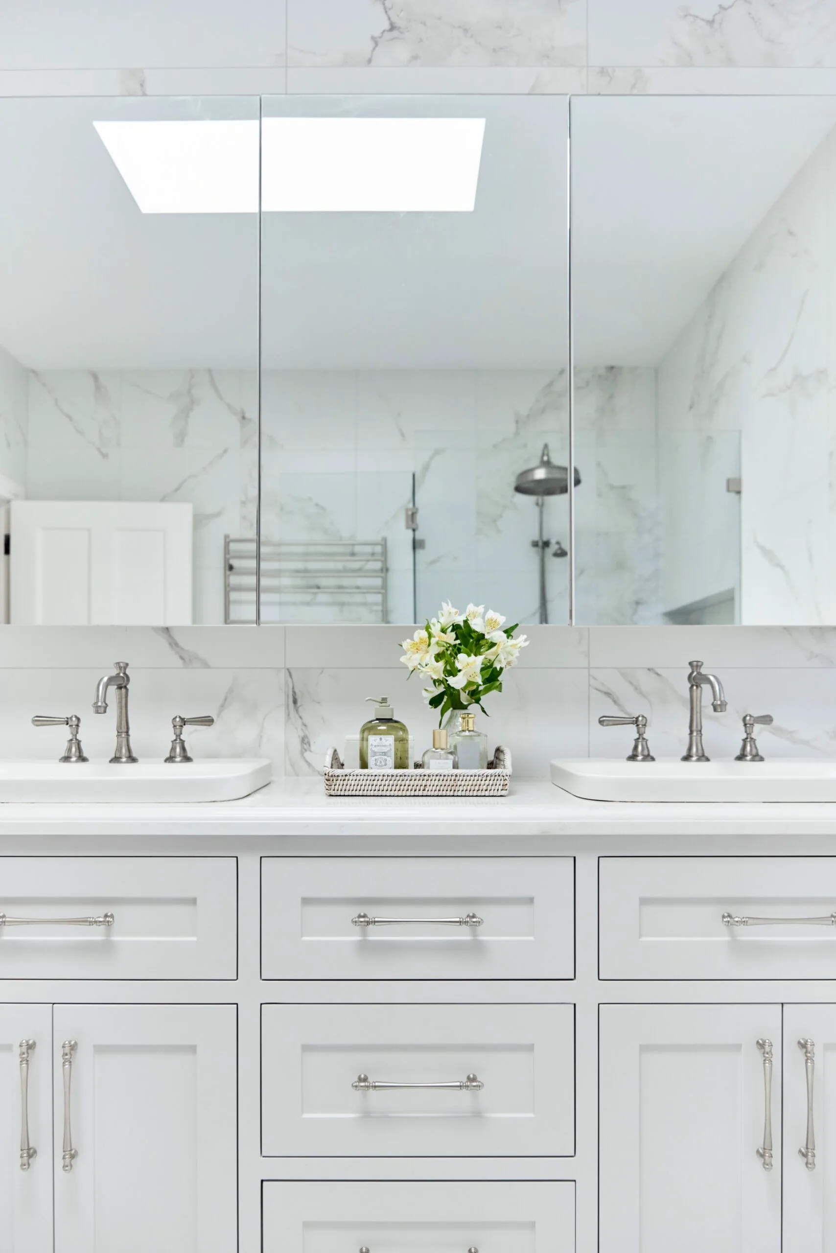 White bathroom with double sinks, marble wall tiles, and a tray with flowers and toiletries on the counter. The vanity, positioned underneath a wall-mounted mirror, has shaker style joinery. Hardware and tapware is chrome.