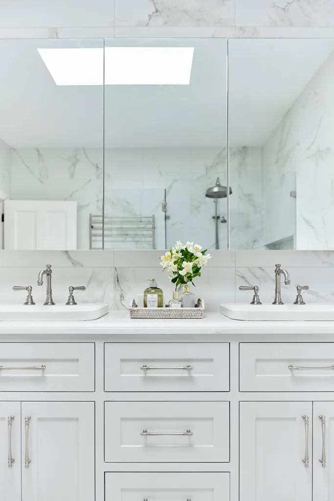 White bathroom with double sinks, marble wall tiles, and a tray with flowers and toiletries on the counter. The vanity, positioned underneath a wall-mounted mirror, has shaker style joinery. Hardware and tapware is chrome.