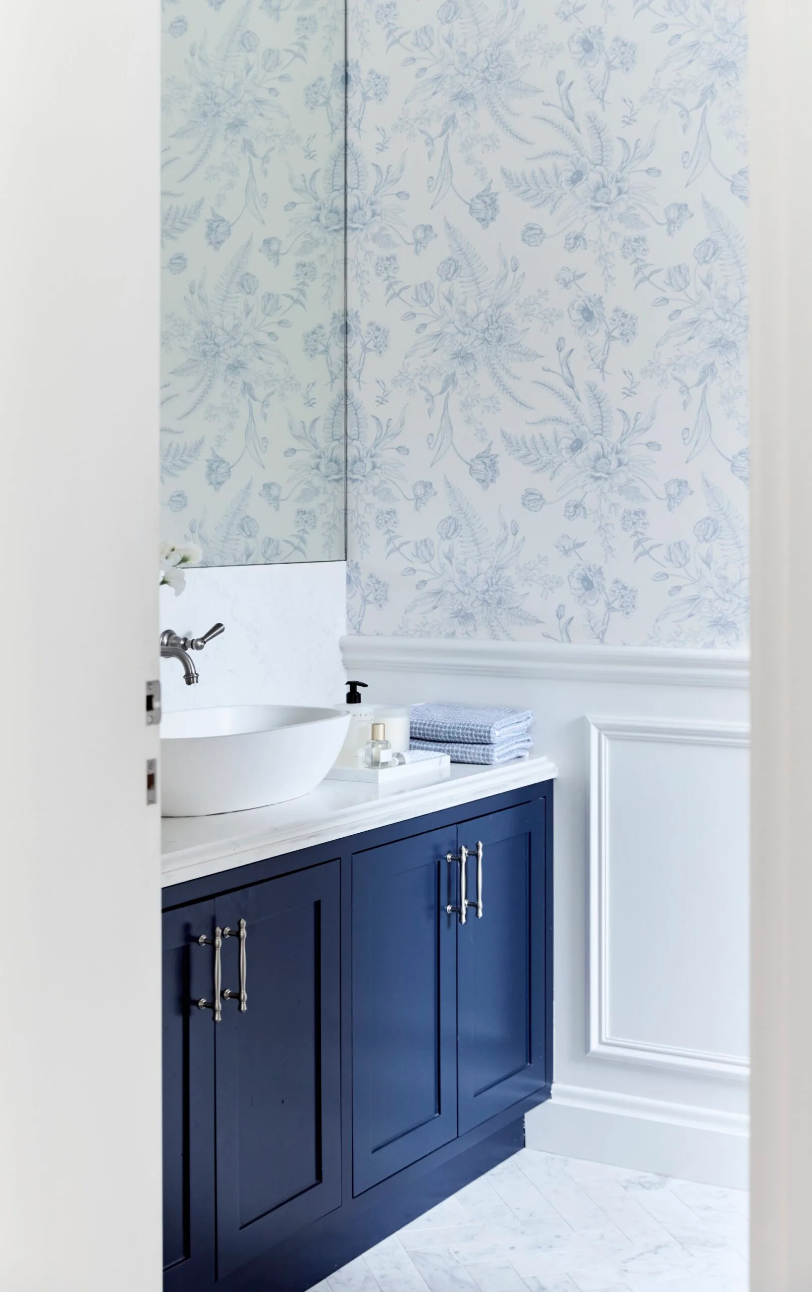 A powder room with blue and white floral wallpaper above white wainscoting, navy cabinets, and a white countertop with a vessel sink.