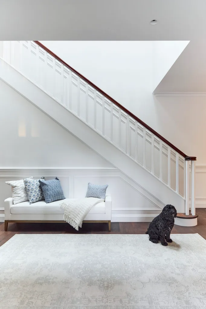 The interior entry of a heritage house. A white sofa with blue cushions is placed against a wall with wainscoting. This wall is under a staircase, with a black cavoodle dog sitting on a light rug, placed atop timber floorboards.