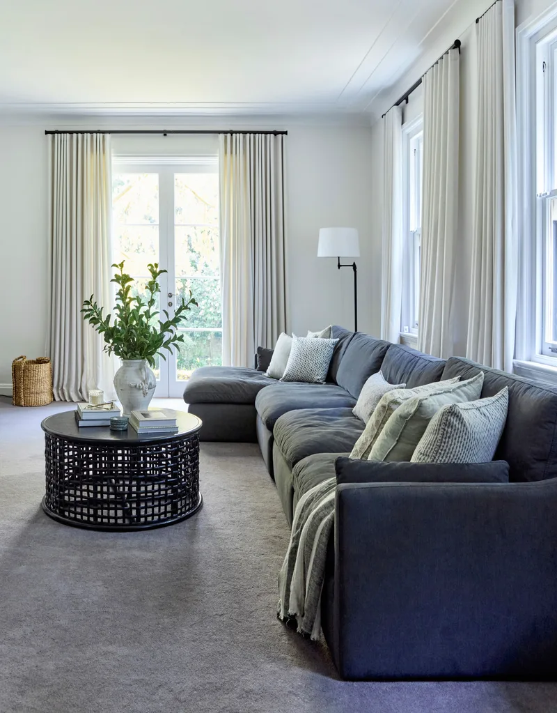 Living room with large gray sectional sofa, round coffee table, floor lamp, and white curtains on tall windows. This is all positioned on lush grey carpet and is in a heritage house.