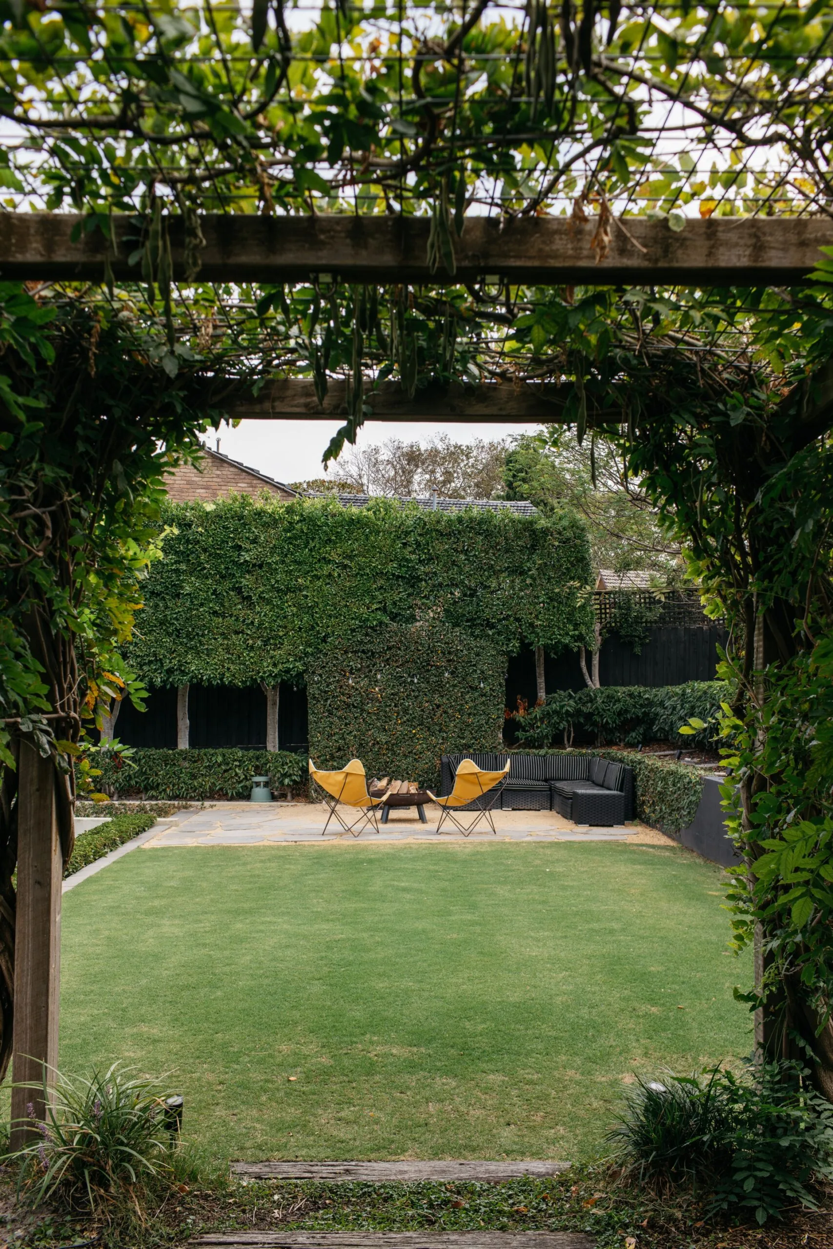 Verdant arbour leading to a garden with lawn, hedges and butterfly chairs.