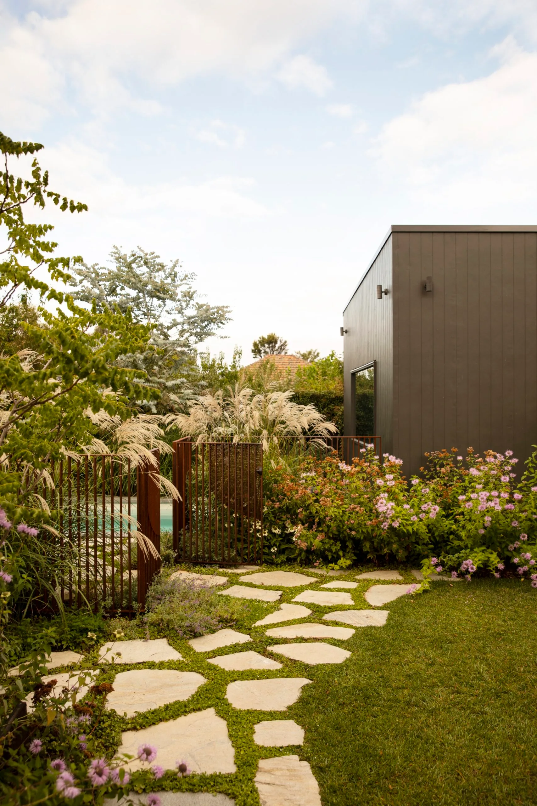 Modern garden with stone paver path, lush plants, and a dark grey building under a bright, cloudy sky. The result of a garden makeover.