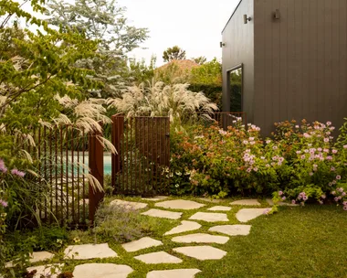 Modern garden with stone paver path, lush plants, and a dark grey building under a bright, cloudy sky. The result of a garden makeover.