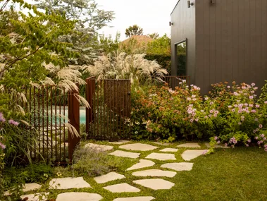 Modern garden with stone paver path, lush plants, and a dark grey building under a bright, cloudy sky. The result of a garden makeover.