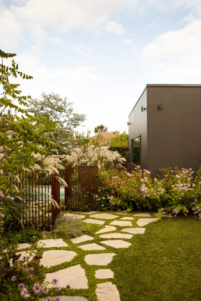 Modern garden with stone paver path, lush plants, and a dark grey building under a bright, cloudy sky. The result of a garden makeover.