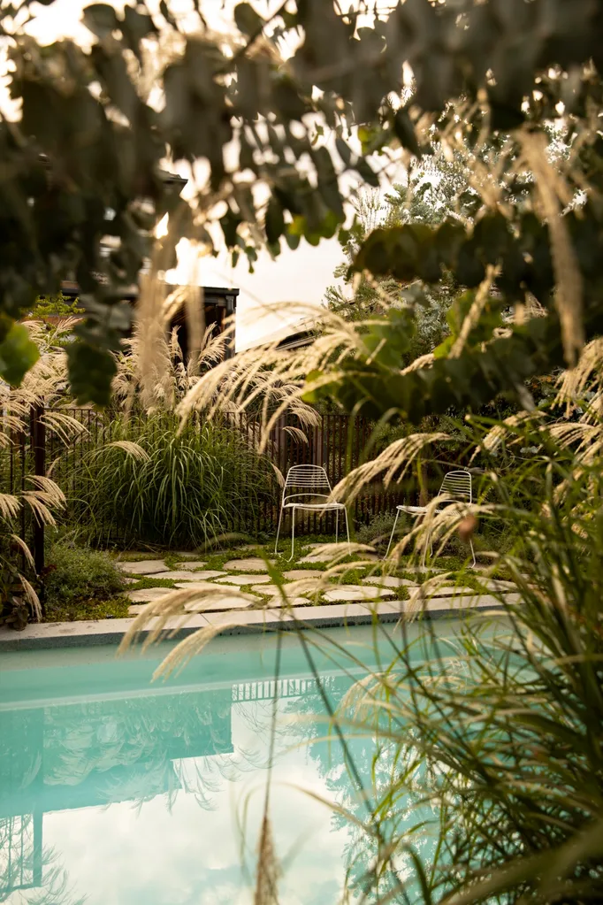 Garden pool with tall grasses, wire chairs, and stone paver path under overcast sky.