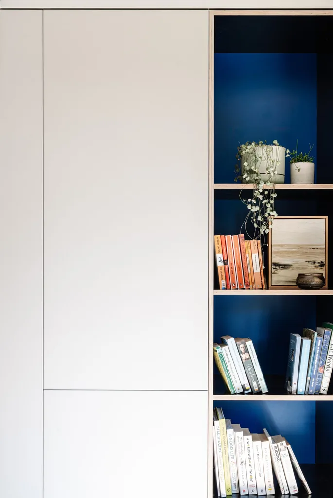 White built-in joinery with four vertical open shelves. The back of the shelves is painted a deep blue and the shelves are decorated with books, artwork and two potted plants.