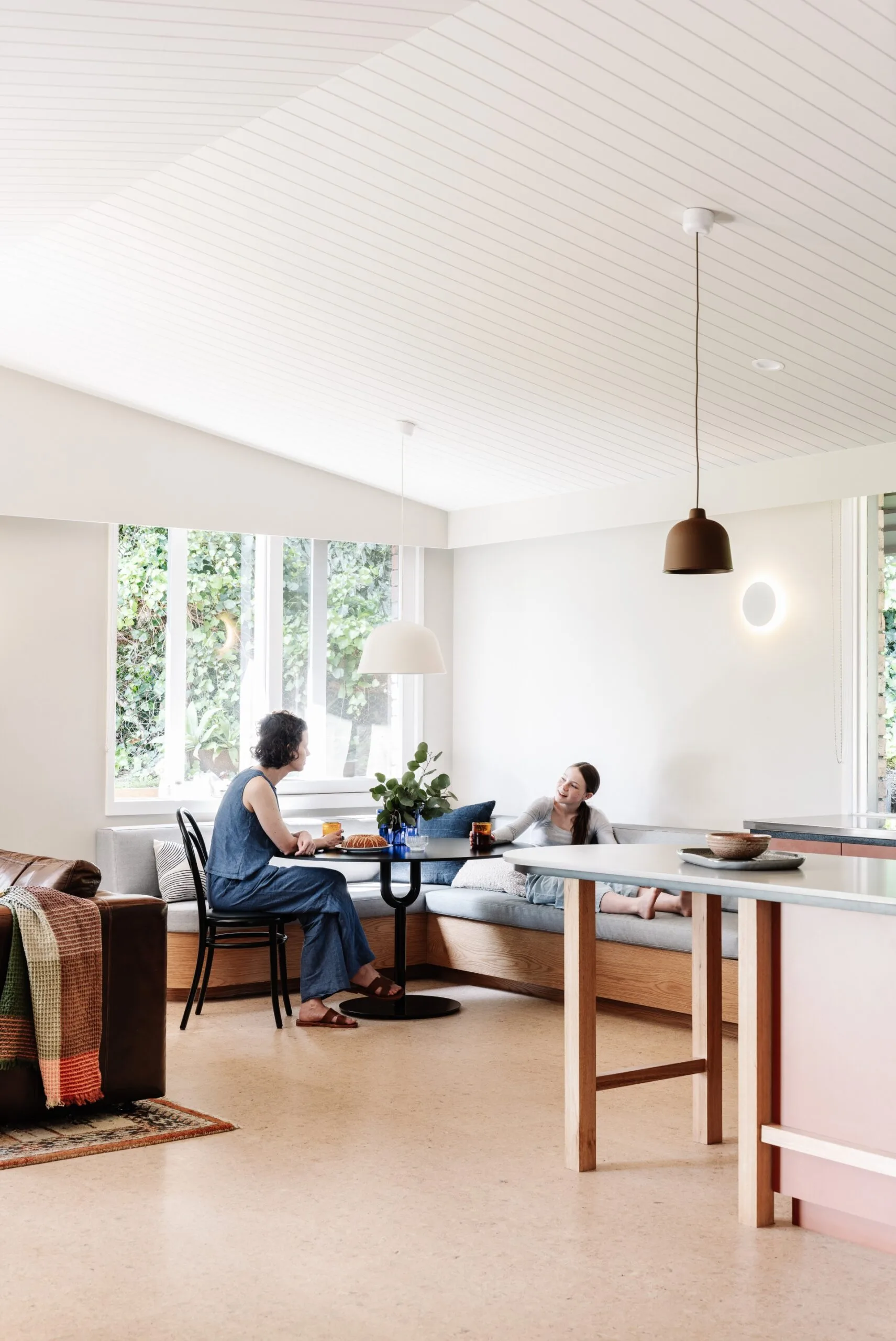 An adult and a child relax and converse at a kitchen table in a bright, modern room with large windows and pendant lights. There is one dining chair set up next to the kitchen table, which the adult sits on. On the other side of the table, in an L-shape, is built-in banquette seating with grey cushions, which the child reclines on.