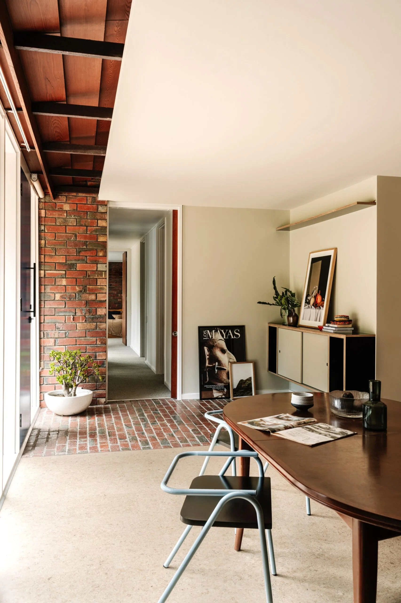 Mid-century style interior with a wooden dining table, black chair, art on a console table, and a potted plant in a bowl. The entrance of the house features a brick wall and brick flooring. This changes to concrete as you step further inside the home