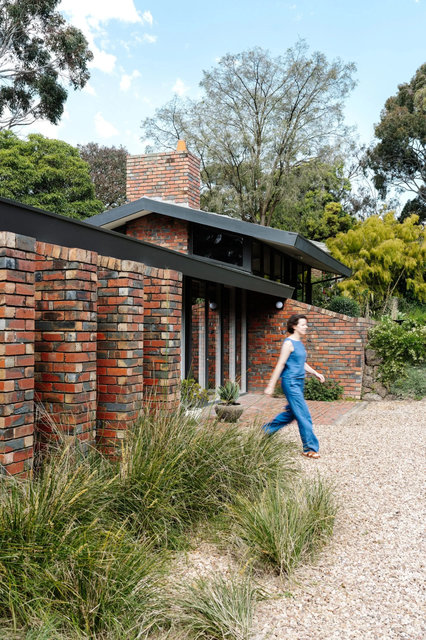 Single-story house with brick accents set in a green, tree-filled landscape. A woman in a blue jumpsuit walks out the home.