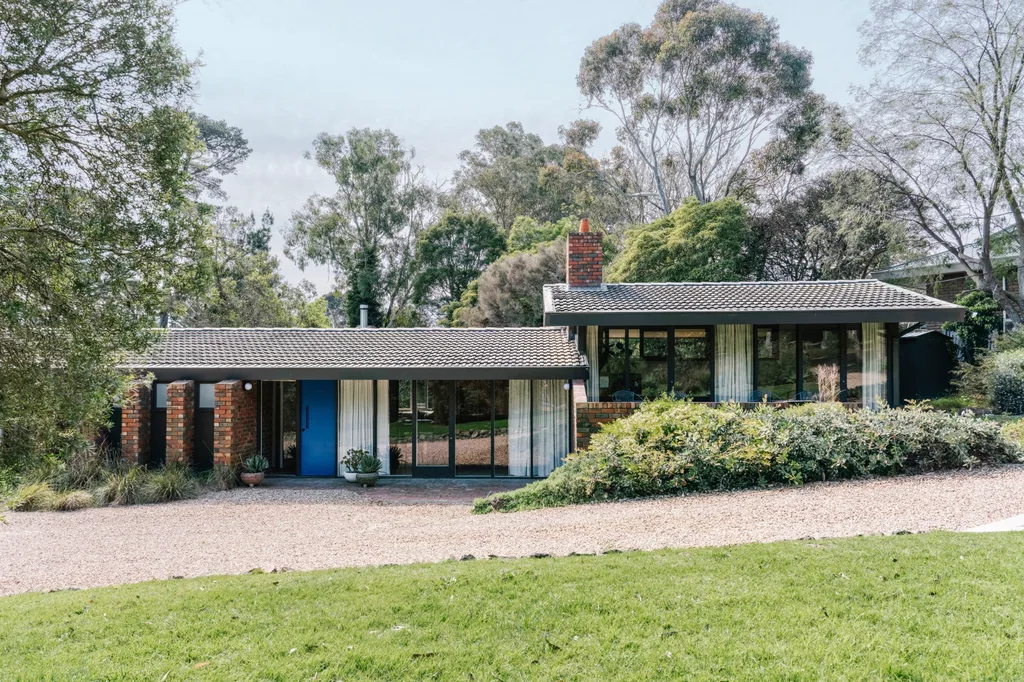 Single-story house with brick accents and a blue front door, set in a green, tree-filled landscape