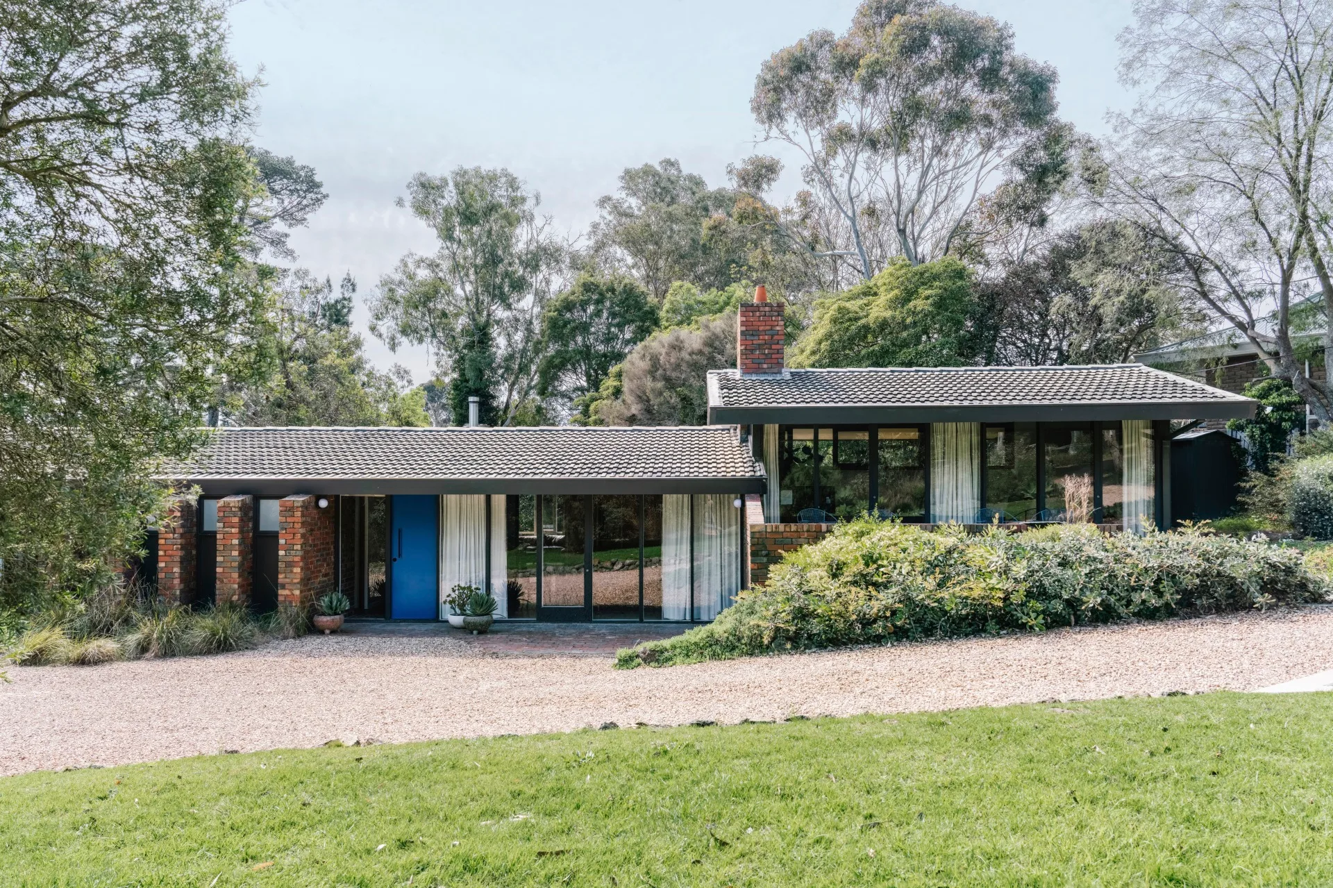 Single-story house with brick accents and a blue front door, set in a green, tree-filled landscape
