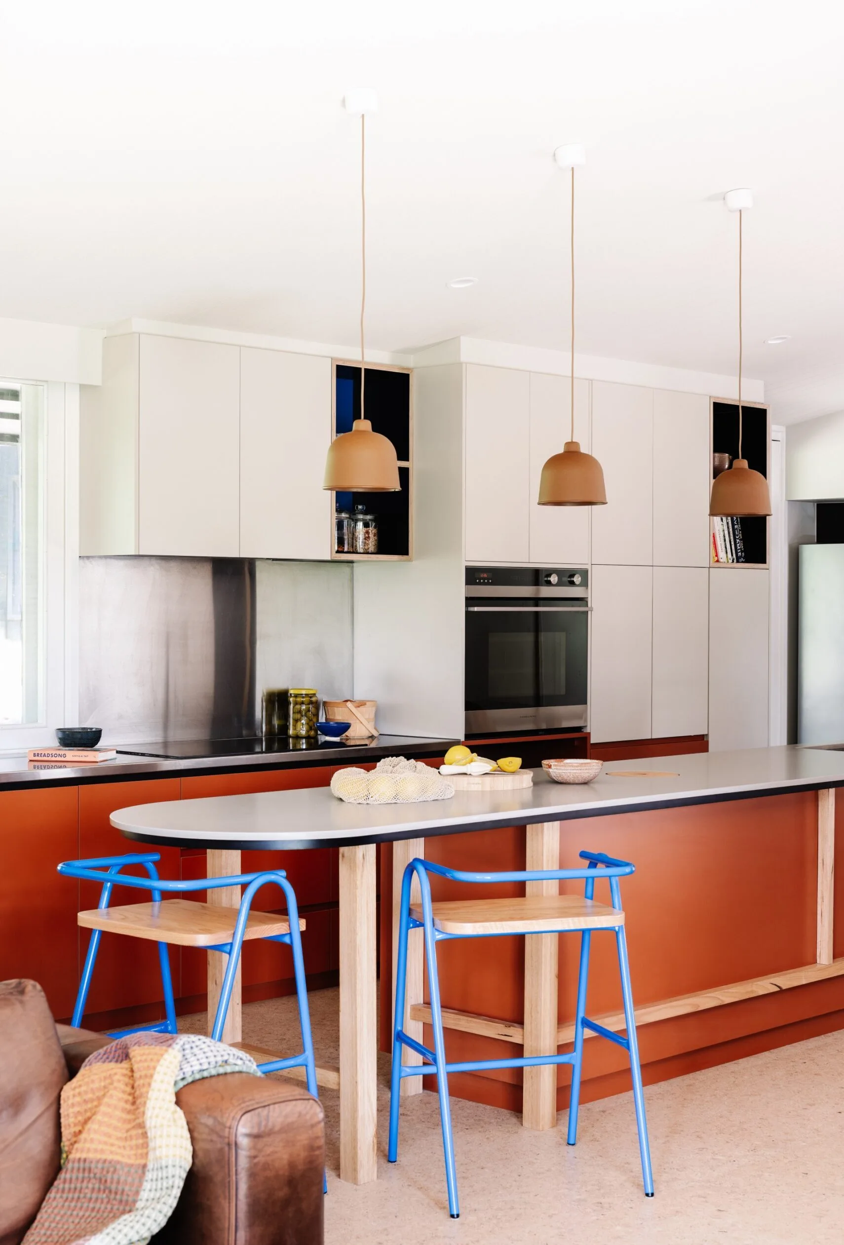 Mid-century style open-plan kitchen with orange island, blue stools, pendant lights and white cabinetry.