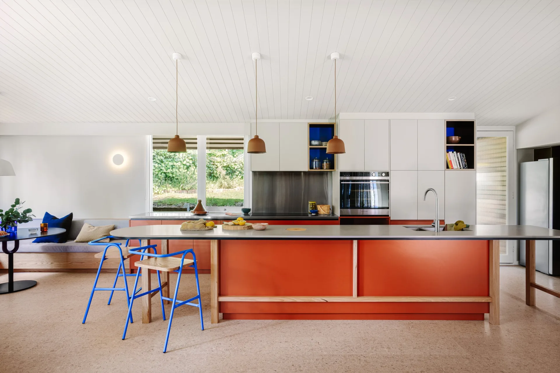 Mid-century style open-plan kitchen with orange island, blue stools, pendant lights, and large windows overlooking greenery. A casual dining nook is to the left of the kitchen with banquette seating and a round dining table.