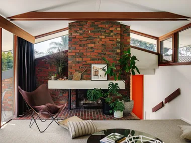 Mid-century style living room with red brick fireplace, brown chair, plants, and a large window with trees outside. Grey carpet is in front of the brick with a round coffee table and scattered cushions around.