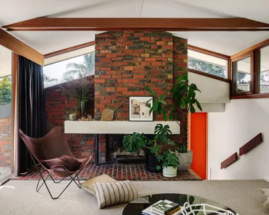 Mid-century style living room with red brick fireplace, brown chair, plants, and a large window with trees outside. Grey carpet is in front of the brick with a round coffee table and scattered cushions around.