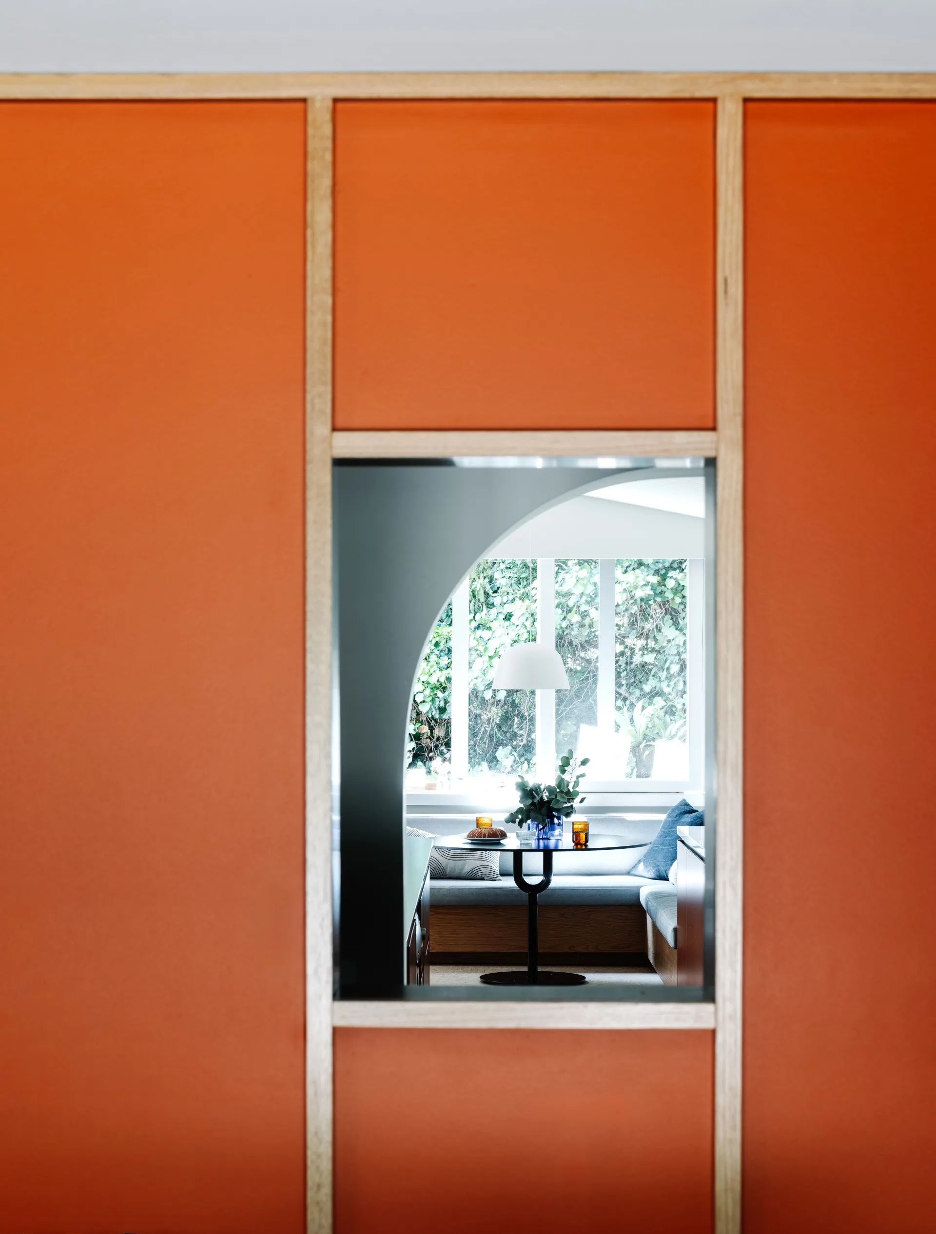 A wall painted orange with wooden panels, with a rectangular window positioned in the middle. This looks through the wall to a casual dining nook with built in banquette seating.