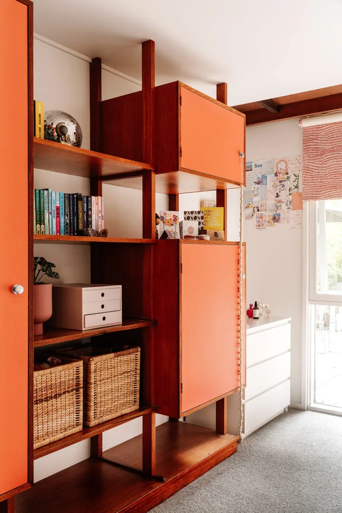 Mid-century modern shelving unit with orange panels, books, woven baskets, and decor items in a bright room with white walls.