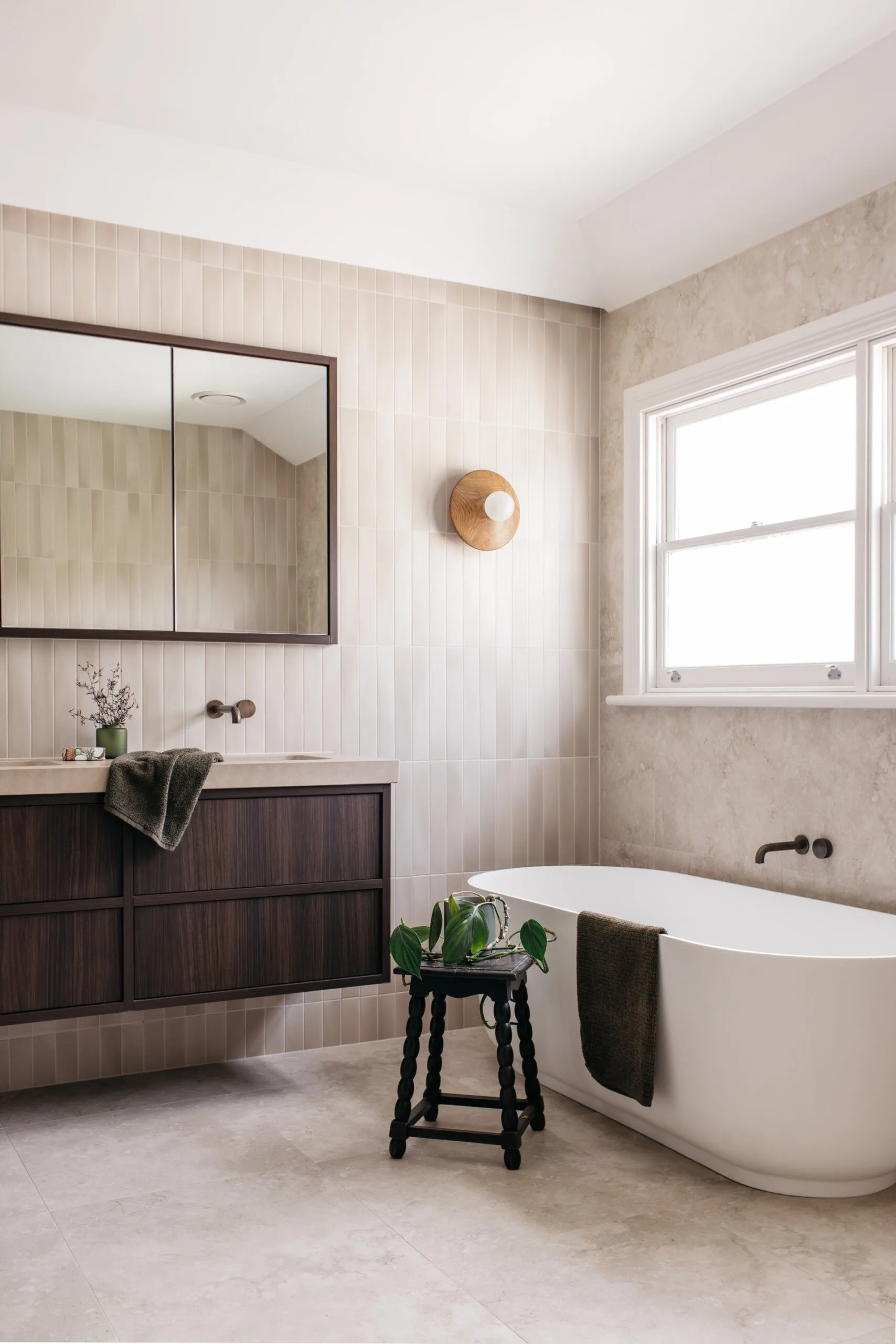 Warm, neutral bathroom featuring a walnut vanity, mirrored cabinet, freestanding bathtub and a bobbin stool.