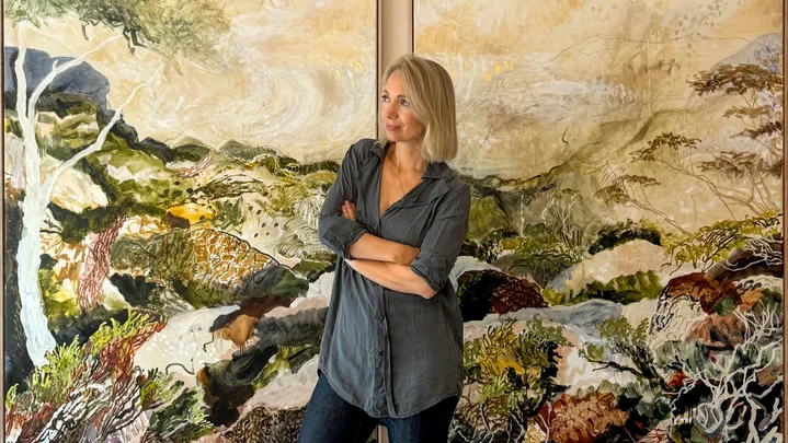 A person in a gray shirt stands in front of two colorful landscape paintings, arms crossed, looking to the side.