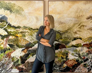 A person in a gray shirt stands in front of two colorful landscape paintings, arms crossed, looking to the side.