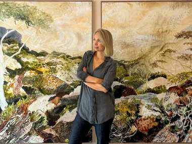 A person in a gray shirt stands in front of two colorful landscape paintings, arms crossed, looking to the side.