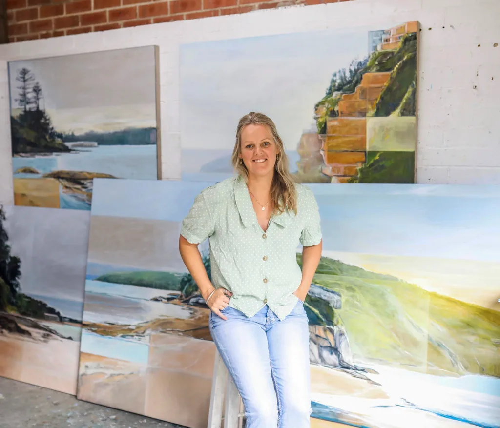 Woman in a light green shirt and jeans stands in front of large landscape paintings in an art studio