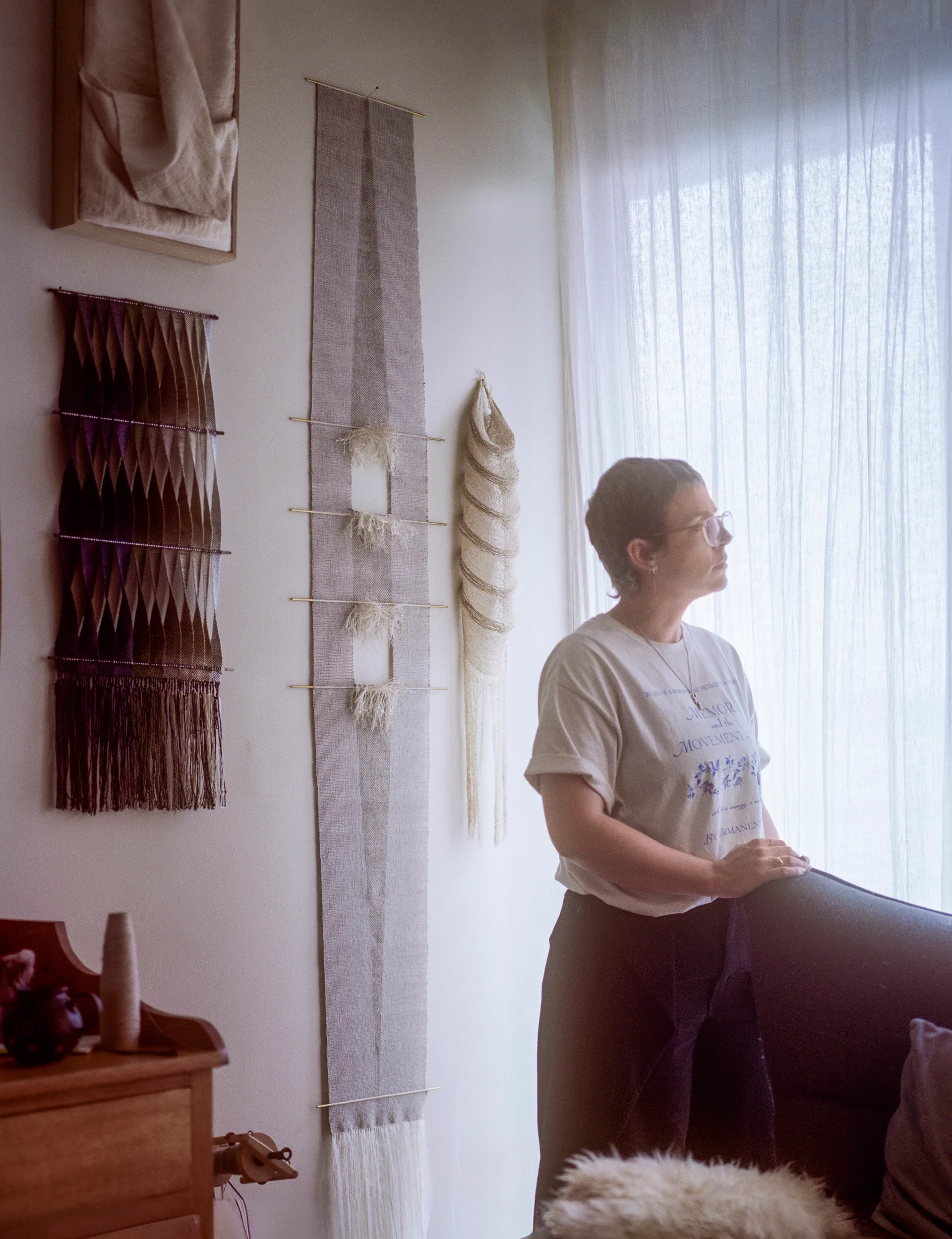 Person gazing out window in a room with woven wall hangings and soft lighting.