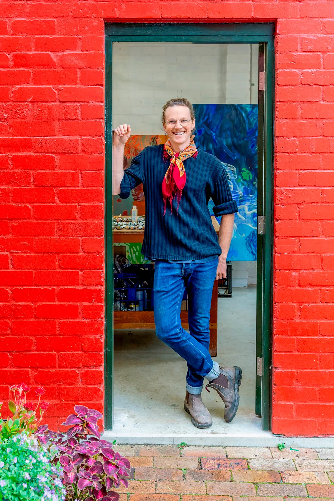 Person standing in a doorway with a red wall, wearing a colorful scarf and casual clothes, smiling warmly. Paintings are visible through the open doorway.