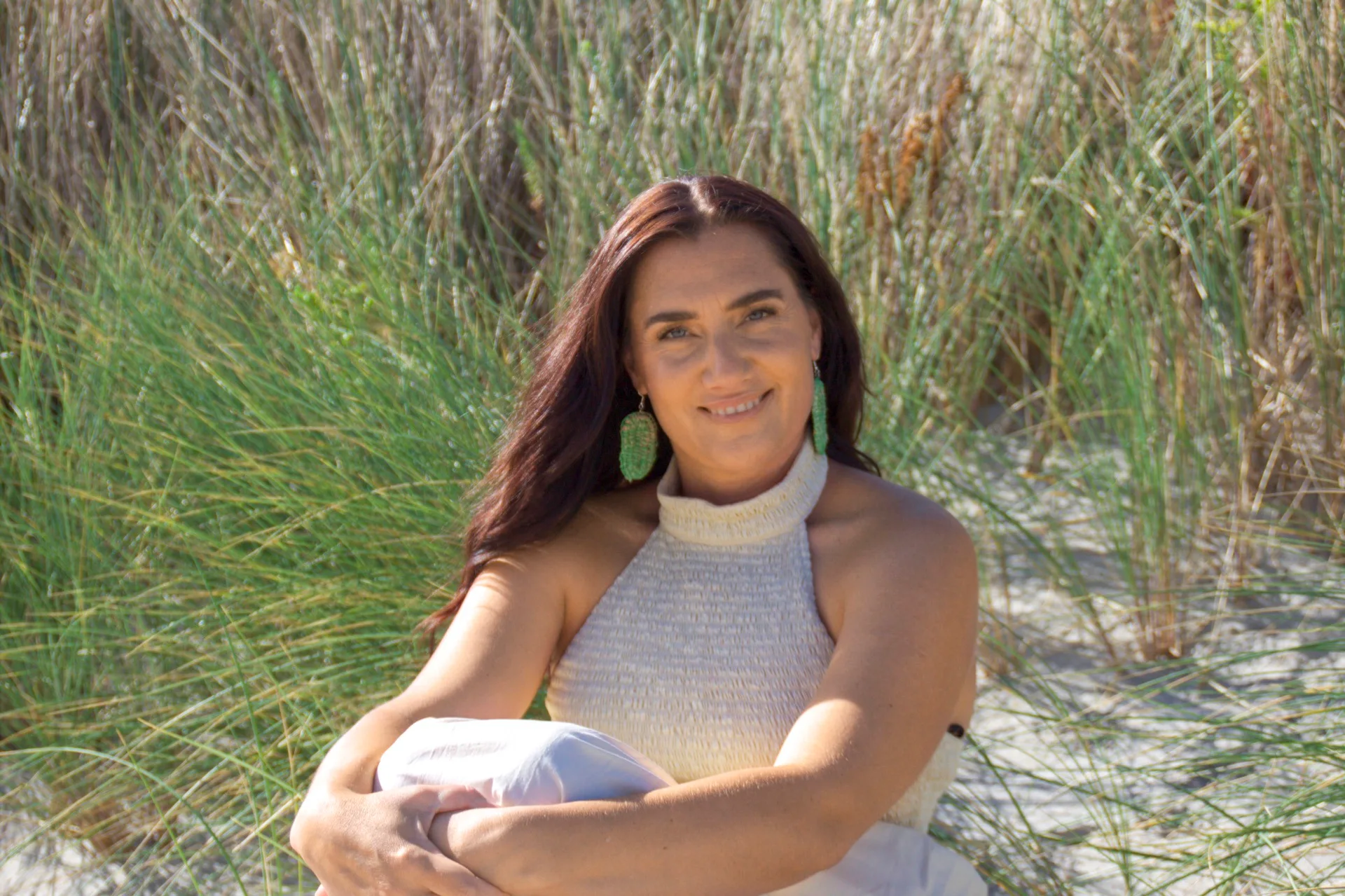 Woman sitting on sand with tall grass, wearing a sleeveless top and green earrings, smiling at the camera.