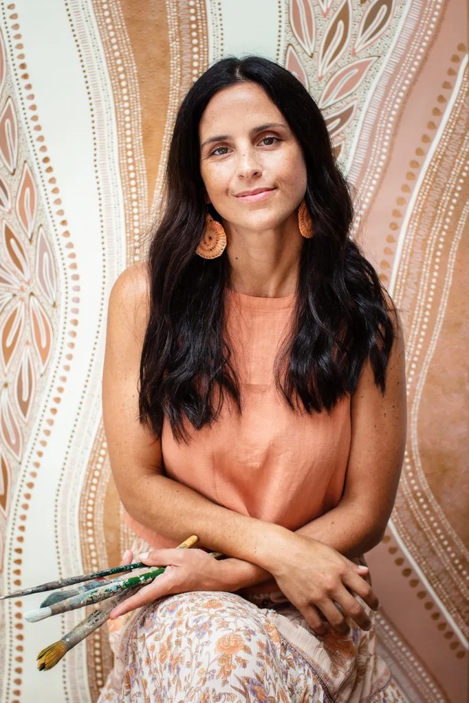 A woman in an orange top holding paintbrushes, seated in front of a a large painting.